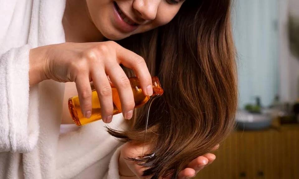 A woman applies argan oil to her damp hair in front of a mirror during dry winter weather